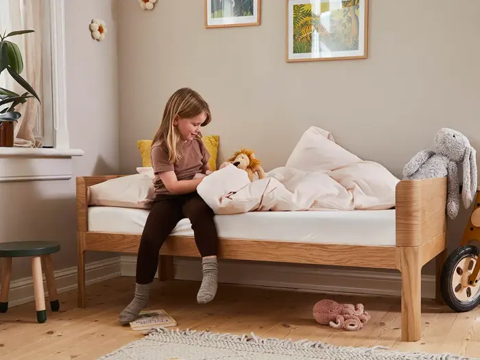 En ung pige sidder på en træseng med lyst sengetøj og holder et tøjdyr i et hyggeligt, veloplyst soveværelse, der er indrettet med legetøj og indrammet kunst.