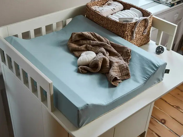 Et puslebord med en blå puslepude, en brun klud, et tøjdyr og en kurv med babyudstyr i et børneværelse med trægulv.