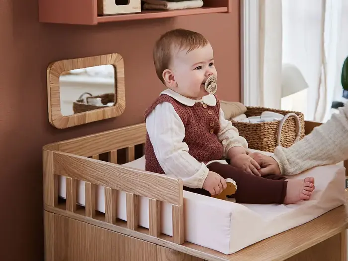 En baby med sut sidder på et puslebord af træ iført en rødbrun vest og en cremefarvet skjorte, mens en voksen hånd forsigtigt holder babyens ben.