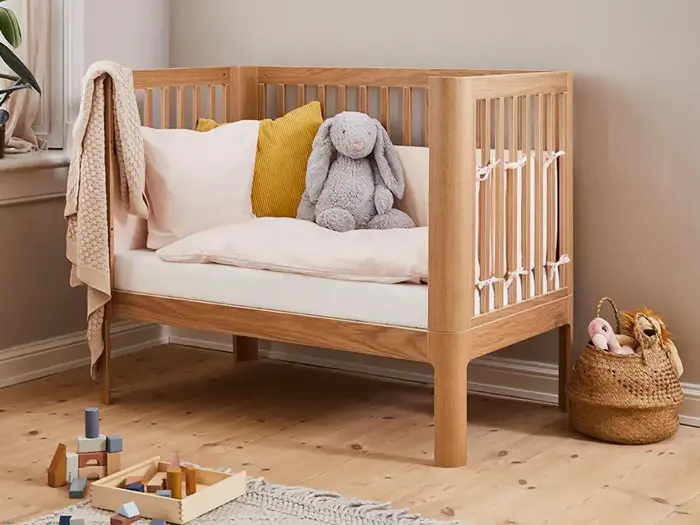 En tremmeseng af træ med lyst sengetøj, en sennepsgul pude og en grå, udstoppet kanin står i et hyggeligt rum med legetøj og en flettet kurv på trægulvet.