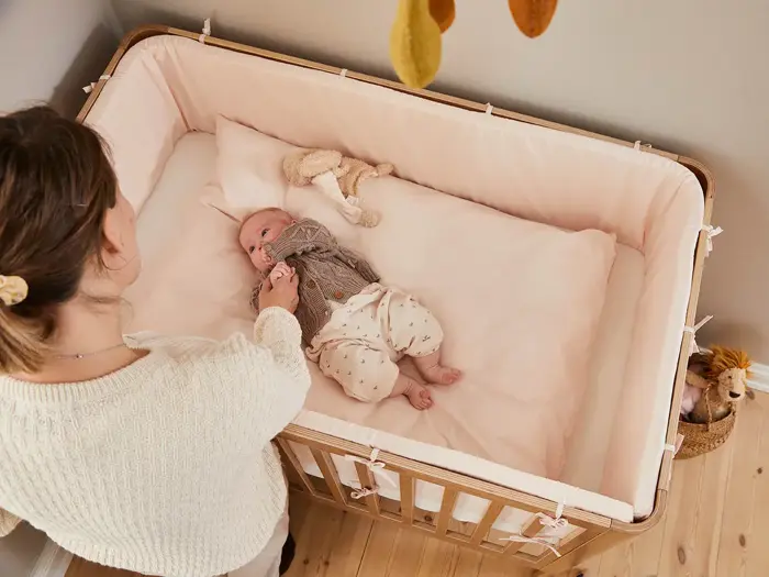 En kvinde står ved siden af en krybbe af træ og holder forsigtigt en baby i hånden, som ligger på et lyserødt lagen med et tøjdyr i nærheden.