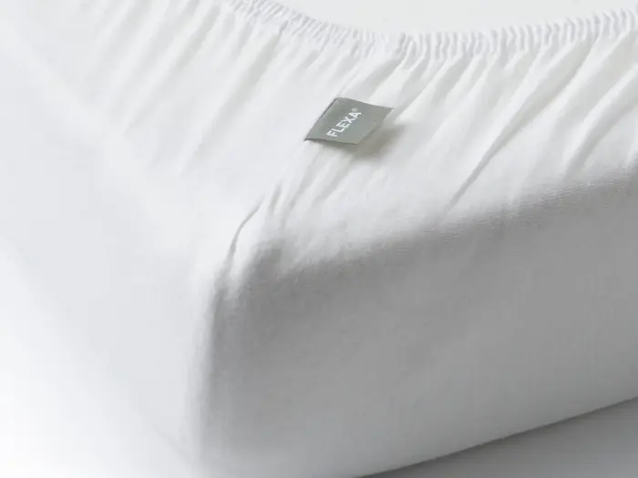Nærbillede af hjørnet af en madras dækket af et hvidt lagen med et gråt mærke med påskriften FLEKKA.