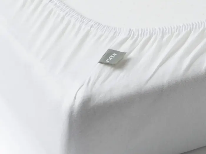 Nærbillede af et hvidt lagen med et gråt mærke med påskriften FLEXA, der er spændt ud over hjørnet af en madras.