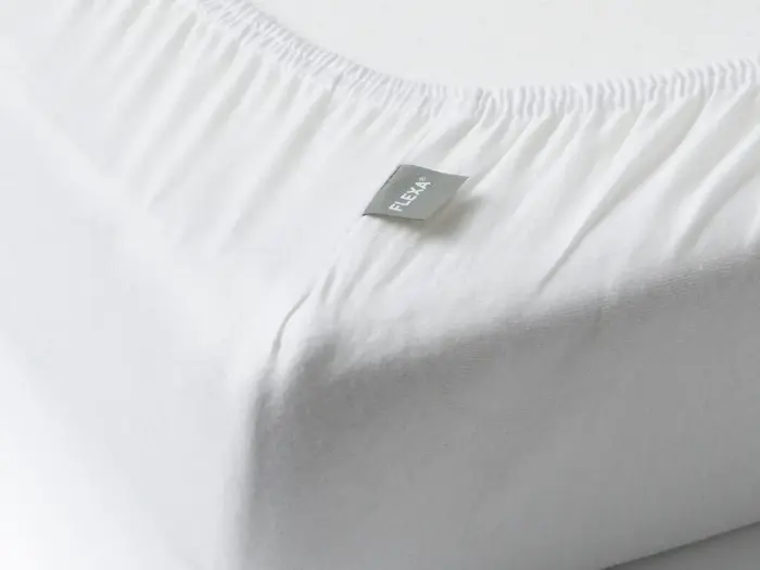 Nærbillede af et hvidt lagen med elastiske kanter på en madras, med et lille gråt mærke med påskriften FLEXA.
