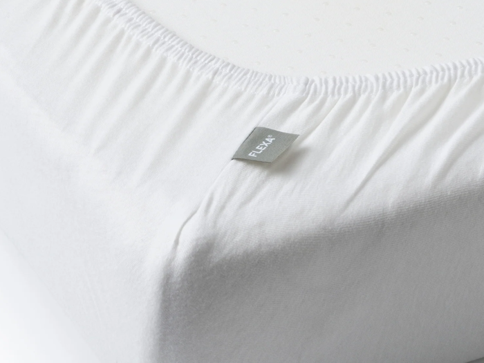 Nærbillede af et hvidt lagen med et synligt FLEXA-mærke, der pænt dækker hjørnet af en madras.
