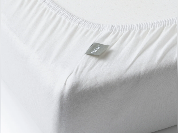 Nærbillede af et hvidt lagen, der pænt dækker hjørnet af en madras, med et lille gråt mærke mærket FLEXA fastgjort til sømmen.