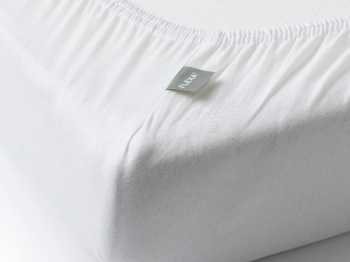 Nærbillede af et hvidt lagen, der dækker en madras, med et lille gråt mærke med påskriften FLEXA i sømmen.