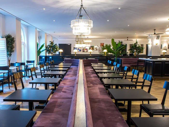 En moderne restaurant med lilla polstrede siddepladser, sorte borde og stole, store lysekroner, trægulv og grønne potteplanter. Rummet er godt oplyst og tomt.