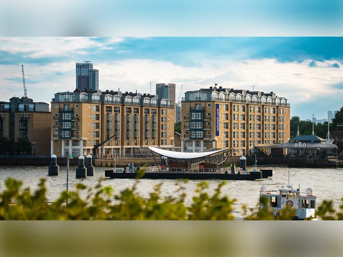 En moderne bygning ved floden står bag en lille dok på vandet, med bybygninger og grønne områder i baggrunden under en delvis overskyet himmel.