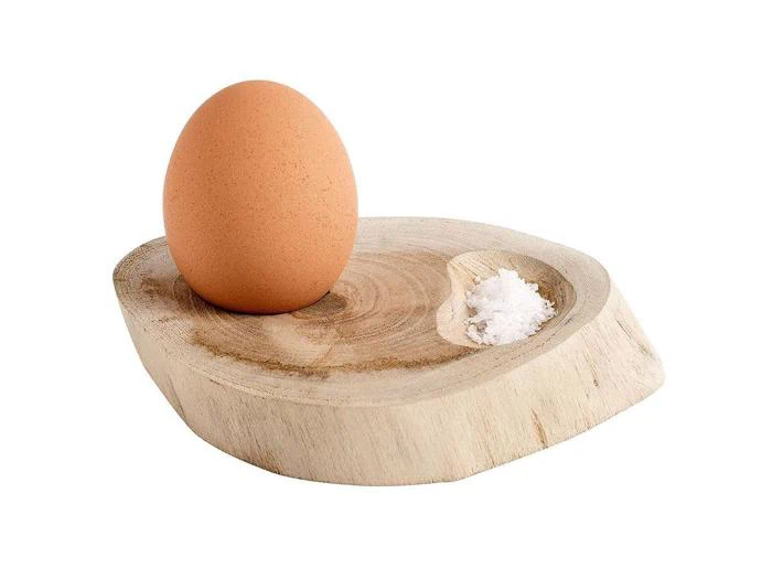 Et brunt æg og en lille bunke salt ligger på et rundt serveringsfad af træ med en udskåret fordybning til hver genstand på en hvid baggrund.