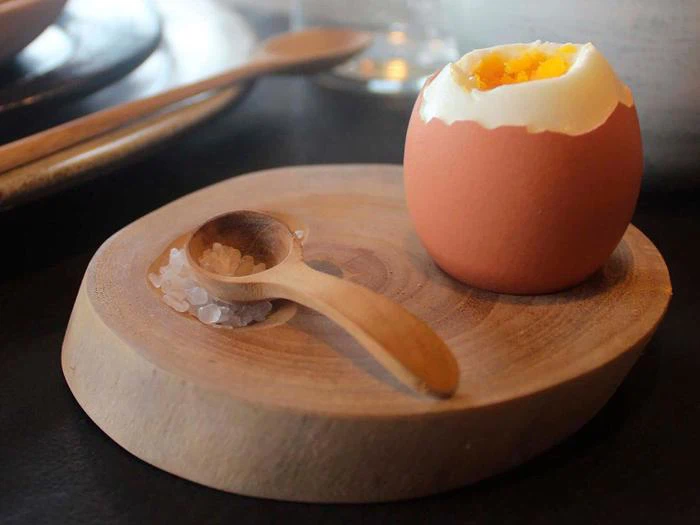 Et blødkogt æg med toppen slået op ligger på en rund træplade ved siden af en træske, der hviler på en lille bunke groft salt.
