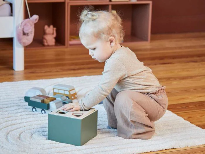 Et lille barn med krøllet blondt hår leger på et hvidt tæppe og lægger figurer i en formsorteringskasse. Legetøjsbiler står i nærheden på trægulvet.