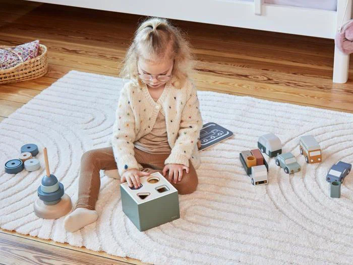 Et lille barn med briller sidder på et hvidt tæppe og leger med et formsorteringslegetøj. Andet trælegetøj og klodser ligger spredt rundt omkring på gulvet.