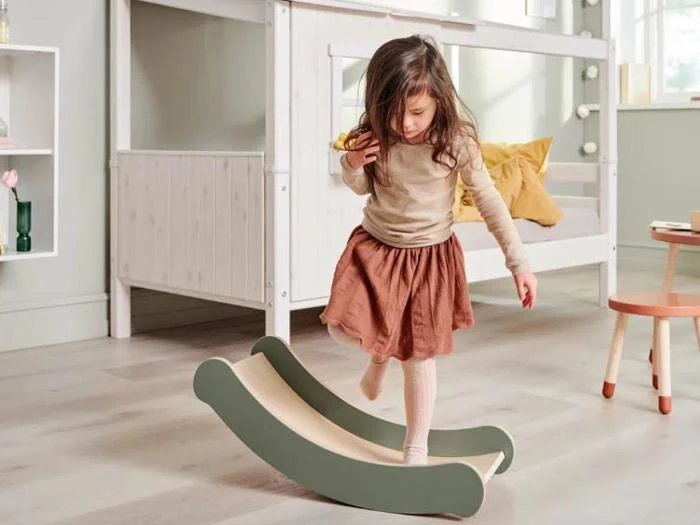 En ung pige balancerer på en buet trægynge i et hyggeligt, lyst soveværelse med en seng og legetøj i baggrunden.