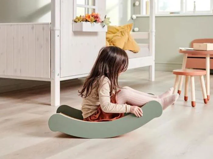 En ung pige sidder på et grønt balancebræt af træ i et hyggeligt, lyst rum med en seng og et lille bord i baggrunden.