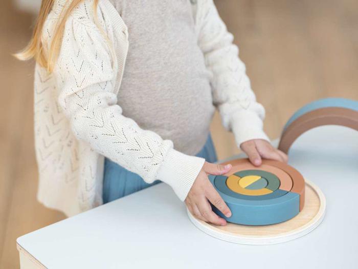 Et barn arrangerer farverige træringe på et hvidt bord iført en lys sweater og en blå nederdel.