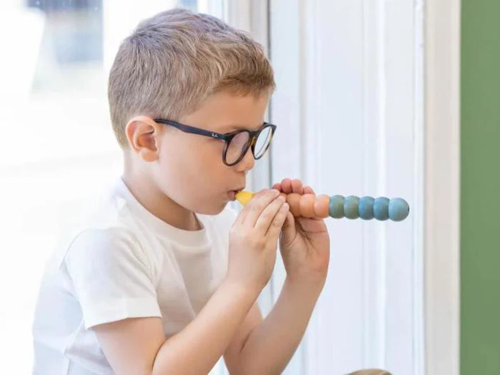 En ung dreng med briller puster ind i et farverigt stykke trælegetøj, mens han sidder ved et vindue og er iført en hvid t-shirt.