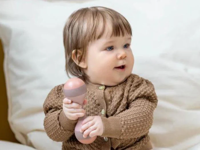 En baby med lysebrunt hår, iført en brun strikket sweater, sidder på en seng med en lyserød og beige rangle i hånden og kigger til siden.