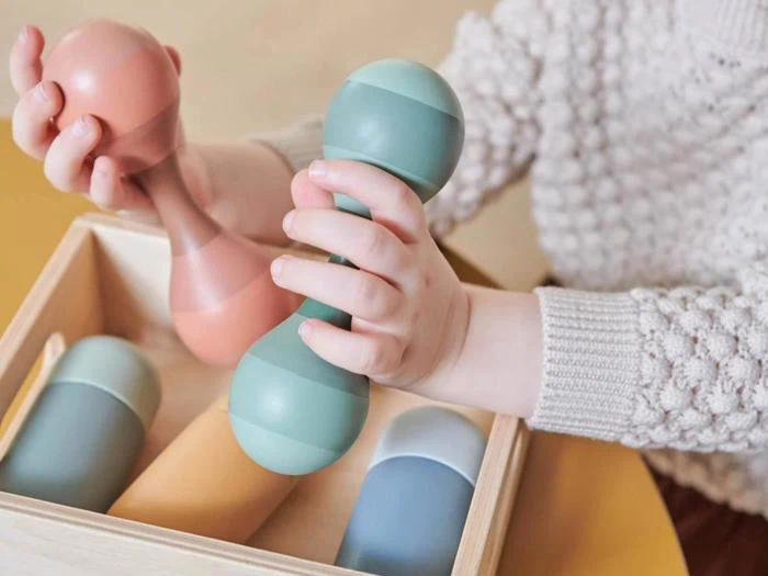 Et barn holder farverige, cylindriske rangler eller legetøj med mere lignende legetøj i en trækasse, iført en strikket sweater.