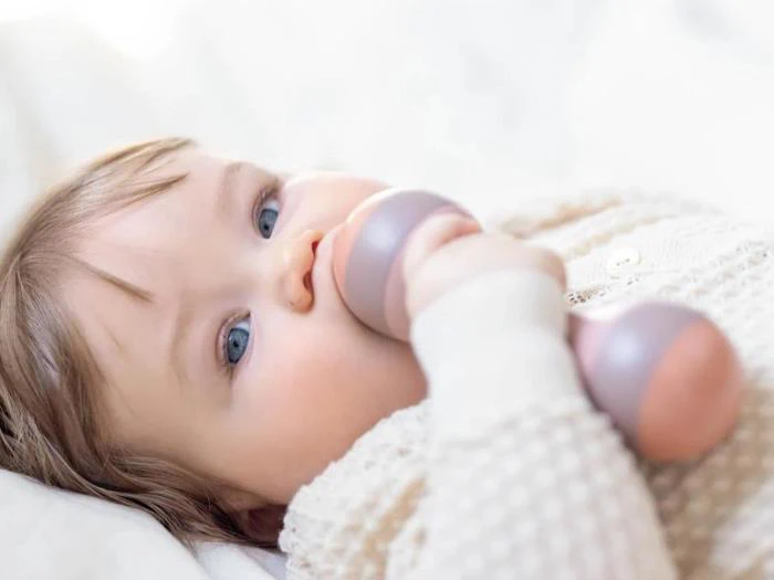 En baby med blå øjne ligger ned og holder og tygger på en blød rangle, mens han er iført en cremefarvet strikket sweater.