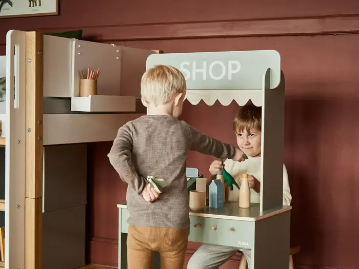 To små børn leger i en butik, hvor den ene giver en genstand til den anden bag en legetøjsdisk med udstillede flasker og beholdere.