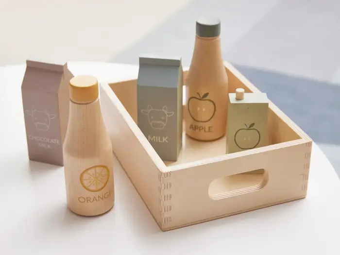 Legetøjsflasker af træ og kartoner mærket chokolademælk, appelsin, mælk, æble og en almindelig karton, arrangeret på et bord med en trækasse.