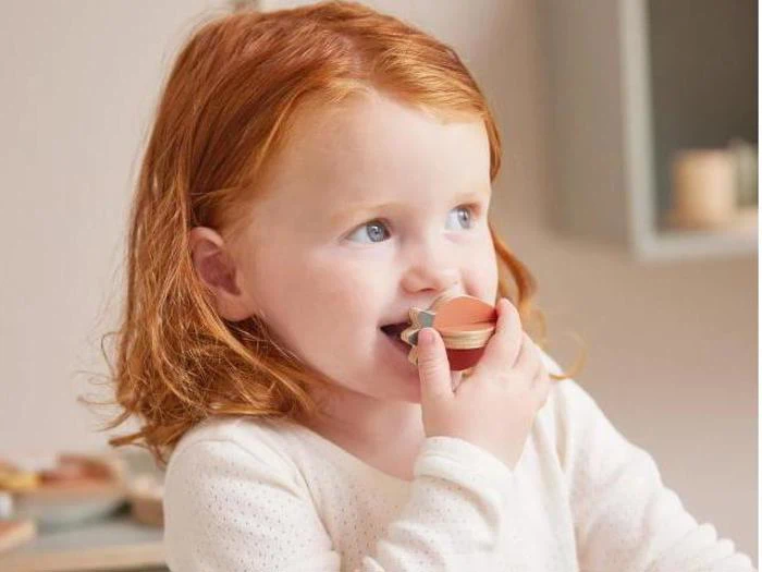 Et lille barn med rødt hår og hvid skjorte holder et lille stykke trælegetøj op til munden og ser nysgerrigt og glad ud.