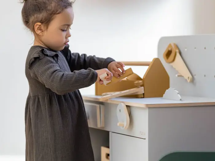 En ung pige i en mørk kjole leger med et legetøjskøkken i træ og lader, som om hun skærer mad ud med en legetøjskniv.
