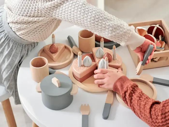 To børn leger ved et hvidt bord med legetøjskage af træ, lys, tallerkener, redskaber og en gryde, der simulerer en fødselsdagsfejring.