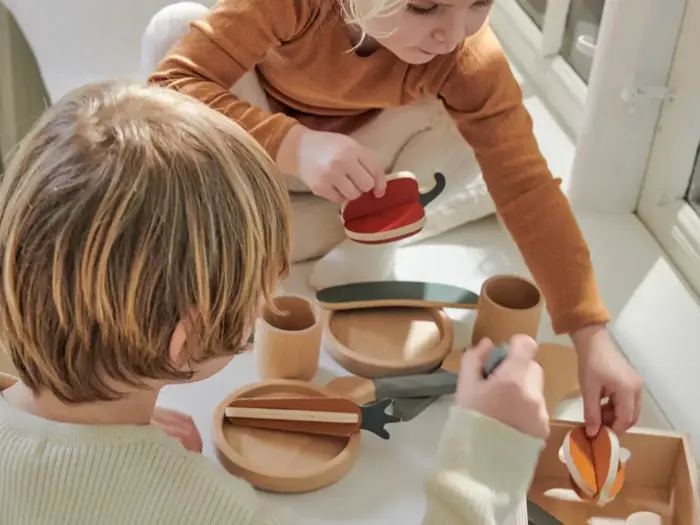 To børn leger med legetøjsmad og -redskaber af træ på et hvidt bord og lader, som om de skærer og serverer stykker i et lyst, solbelyst rum.