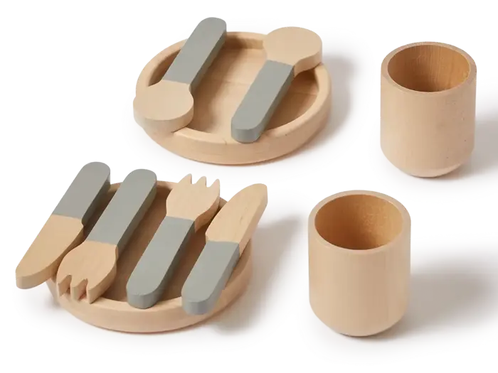 Legetøjsfad af træ med to tallerkener, to kopper og redskaber med gråt skaft, herunder gafler, knive og skeer, arrangeret pænt på en hvid overflade.