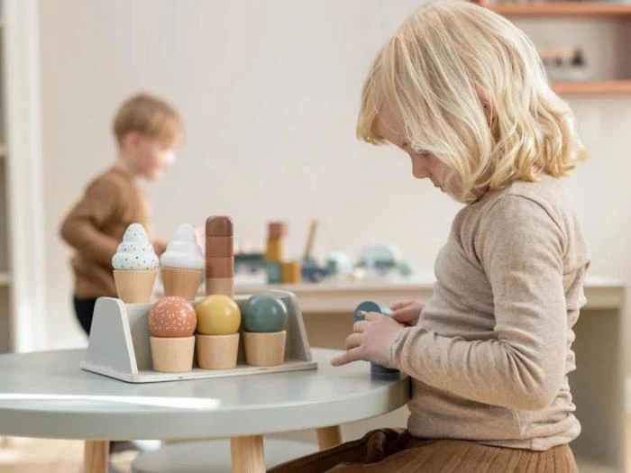 Et lille barn leger med legetøjsis af træ ved et lille bord, mens et andet barn er sløret i baggrunden.