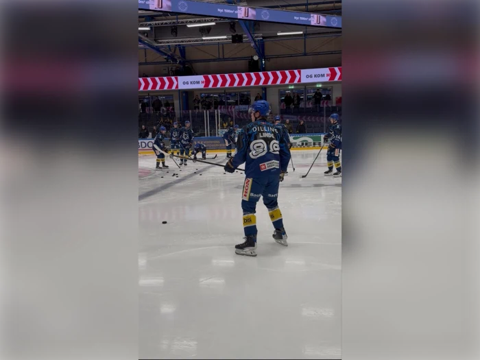 Ishockeyspillere i blå trøjer varmer op på isbanen, og i forgrunden står en spiller med en stav og en puck.