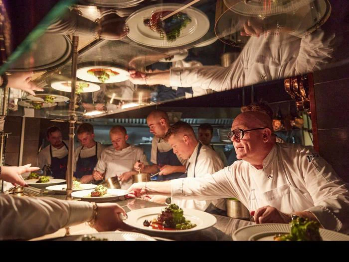 En gruppe kokke i et travlt køkken anretter omhyggeligt retter under varmt lys, mens en kok rækker en tallerken til en tjener.