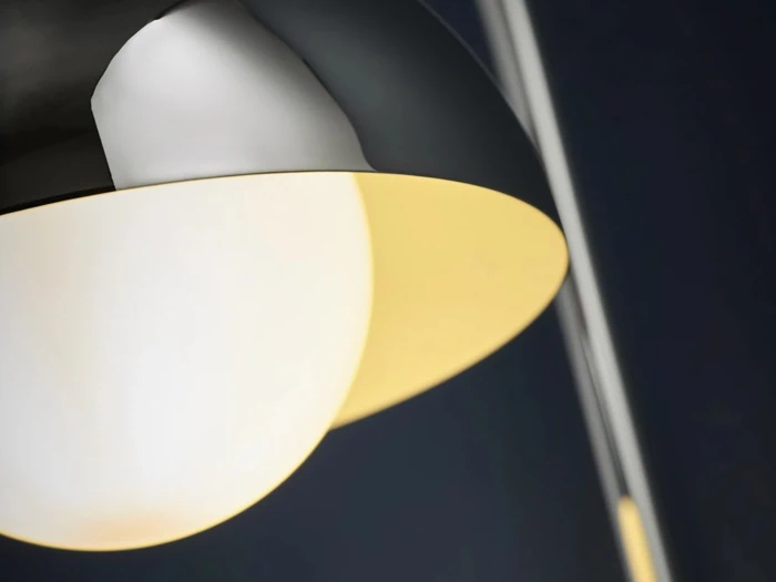 Nærbillede af en moderne lampe med en metalskærm og en lysende hvid pære, fotograferet på en mørk baggrund.