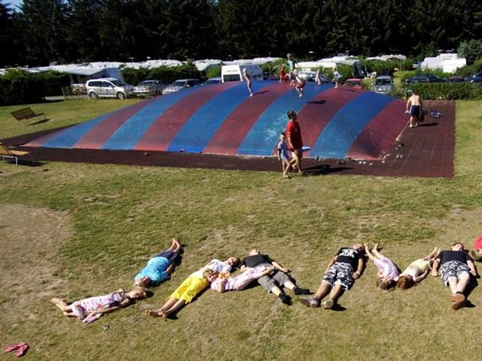 En gruppe mennesker ligger på græsset og staver LAMP med deres kroppe i nærheden af en stor, stribet oppustelig legestruktur og andre mennesker, der leger.