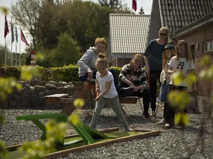 En ung pige spiller minigolf, mens fire personer ser på og smiler bag hende på en solrig dag med bygninger og grønne områder i baggrunden.