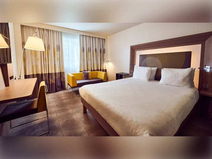 Moderne hotelværelse med en stor seng, hvidt sengetøj, skrivebord og stol, gul sofa, gulvlampe og vindue med mønstrede gardiner.