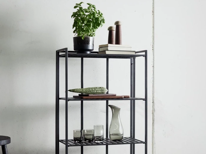 På en sort metalhylde står en potteplante med basilikum, salt- og peberbøsser, bøger, en træbakke med et dekorativt objekt, en keramikkande og tre glaskopper.