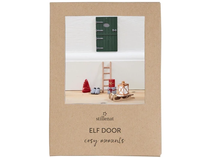 En grøn miniaturedør, stige, rød hat, støvler, lanterne og kælk er arrangeret mod en hvid væg på en træoverflade, udstillet på en brun emballage mærket Elf Door cosy moments.