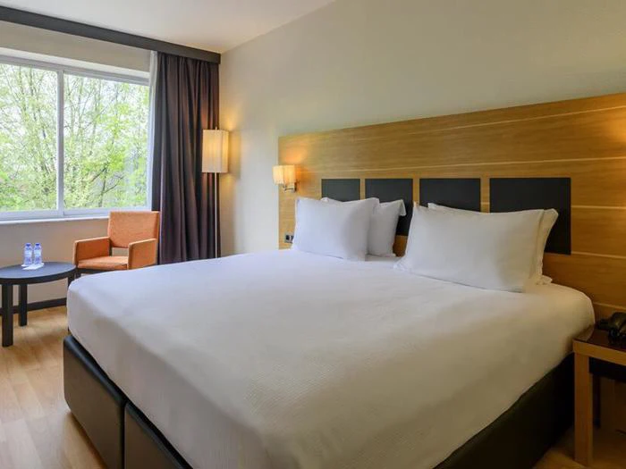 Et moderne hotelværelse med en stor, pænt redt seng, to puder, et hovedgærde af træ, et vindue, et bord med to vandflasker og en orange lænestol.