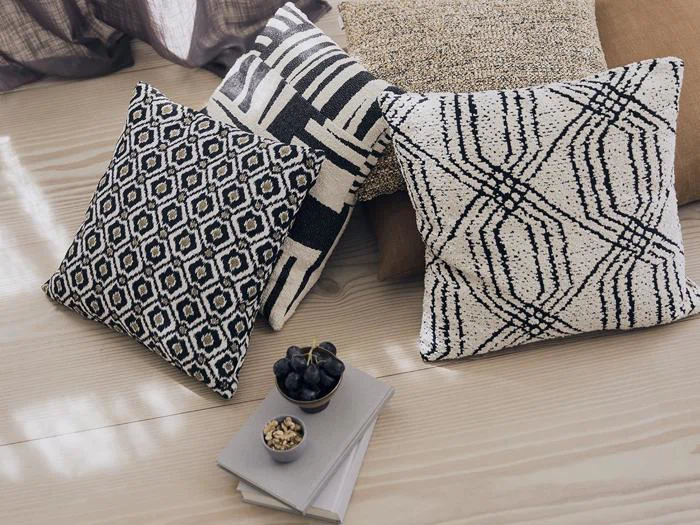 Fire pyntepuder med geometriske mønstre på et lyst trægulv ved siden af to bøger, en lille skål med nødder og en skål med vindruer. Sollyset skaber bløde skygger på gulvet.