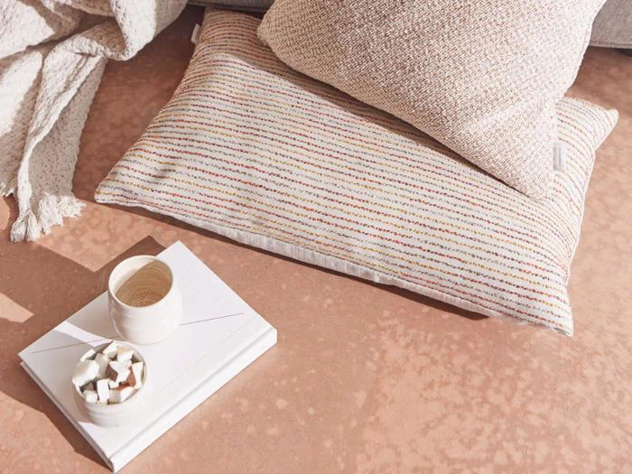 En hyggelig scene med to strukturerede beige puder, en hvid bog, en beige kop og en skål med sukkerknalder på en ferskenfarvet overflade.