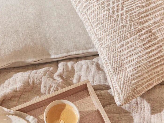 En beige strukturpude, en blød dyne og en træbakke med en kop espresso på en pænt redt seng i neutrale toner.