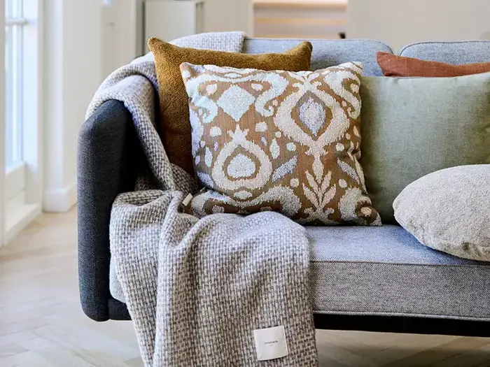 En grå sofa med mønstrede brune og hvide puder, en grøn pude, en beige pude og et foldet tekstureret gråt tæppe, der er draperet over armen.
