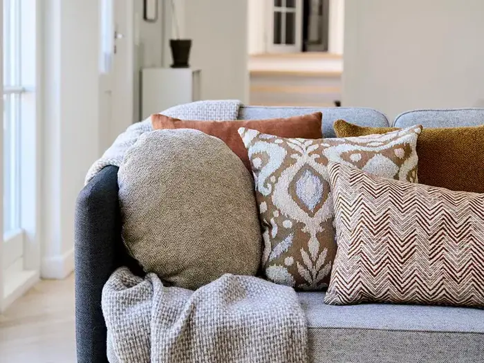 En hyggelig grå sofa med et strikket tæppe og fire pyntepuder i beige, brun og mønstret design i en lys, moderne stue.