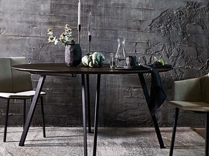 Et moderne spisebord med sorte ben, dækket med en vase med blomster, stearinlys, en glaskaraffel og artiskokker, flankeret af to beige stole mod en struktureret grå væg.