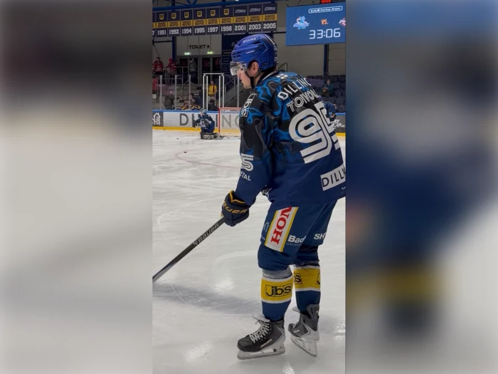 En ishockeyspiller i blåt udstyr med nummer 96 på trøjen glider hen over isen med en ishockeystav i hånden, mens en anden spiller og banden ses i baggrunden.