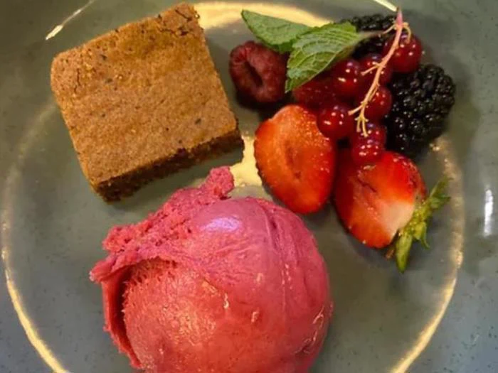 En tallerken med et firkantet stykke kage, en kugle lyserød sorbet og en pynt af friske bær og mynteblade.