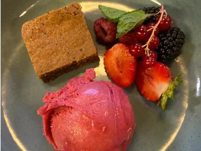 En tallerken med en brownie, en kugle bærsorbet og en blanding af friske bær, herunder jordbær, hindbær, brombær og ribs, pyntet med et mynteblad.